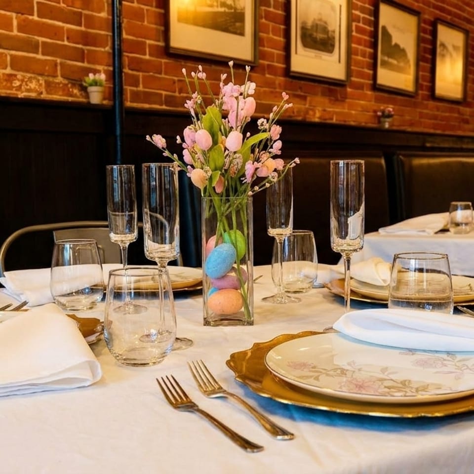 Restaurant dining table set with white tablecloth, floral plates, and a vase of pink flowers and Easter eggs.