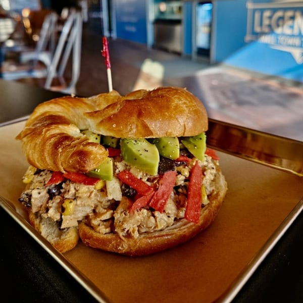 Chicken salad croissant sandwich topped with avocado slices and red tortilla strips on a gold tray.