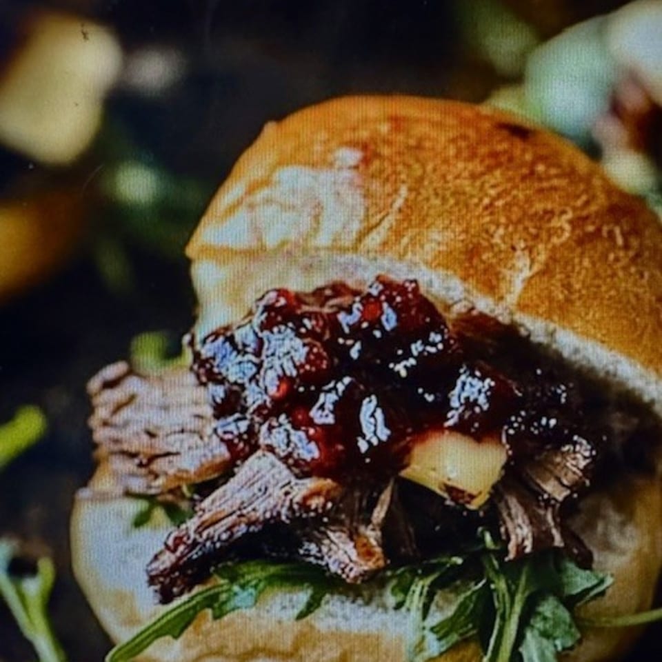 Close-up of a slider with pulled meat, dark jam, cheese, and arugula on a bun.