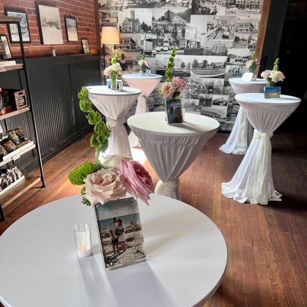 Several white-draped cocktail tables with floral centerpieces and framed photos, against a brick wall and photo collage.