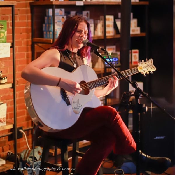 A woman with red hair and a white guitar sings into a microphone on a stage.