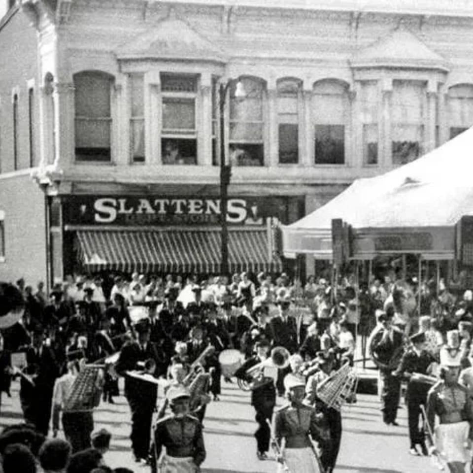 A black and white photo of a marching band parade passing Slattens department store.