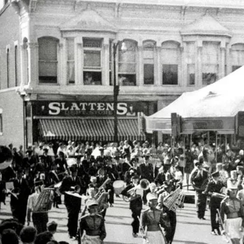 A black and white photo of a marching band parade passing Slattens department store.